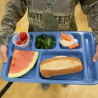 child holding a blue lunch tray with watermelon, carrots, greens, and tomatoes